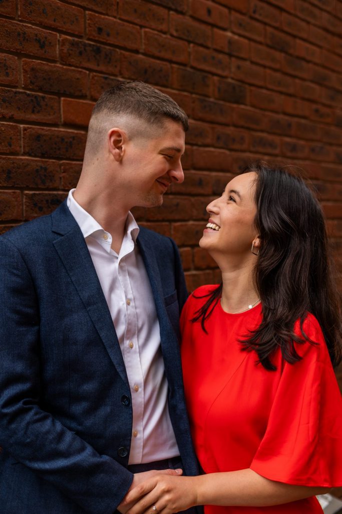 Newly engaged couple walking together in Ancoats, Manchester - The bride to-be is wearing a vivid red dress & the groom to-be is wearing a blue suit - Engagement Shoot - Isabelle B Photography