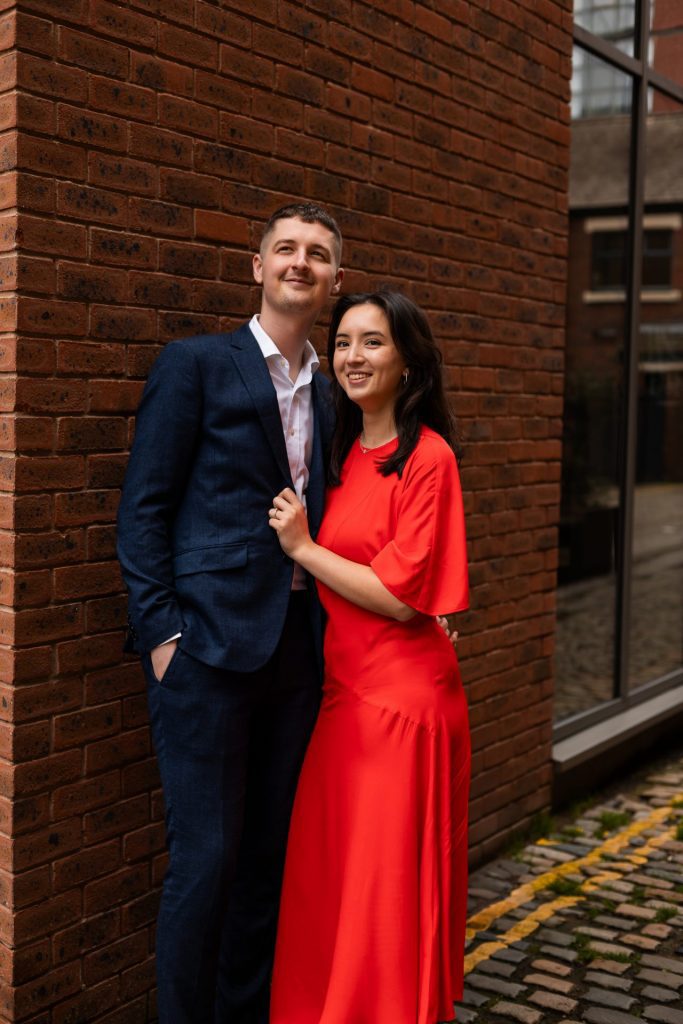 Newly engaged couple walking together in Ancoats, Manchester - The bride to-be is wearing a vivid red dress & the groom to-be is wearing a blue suit - Engagement Shoot - Isabelle B Photography