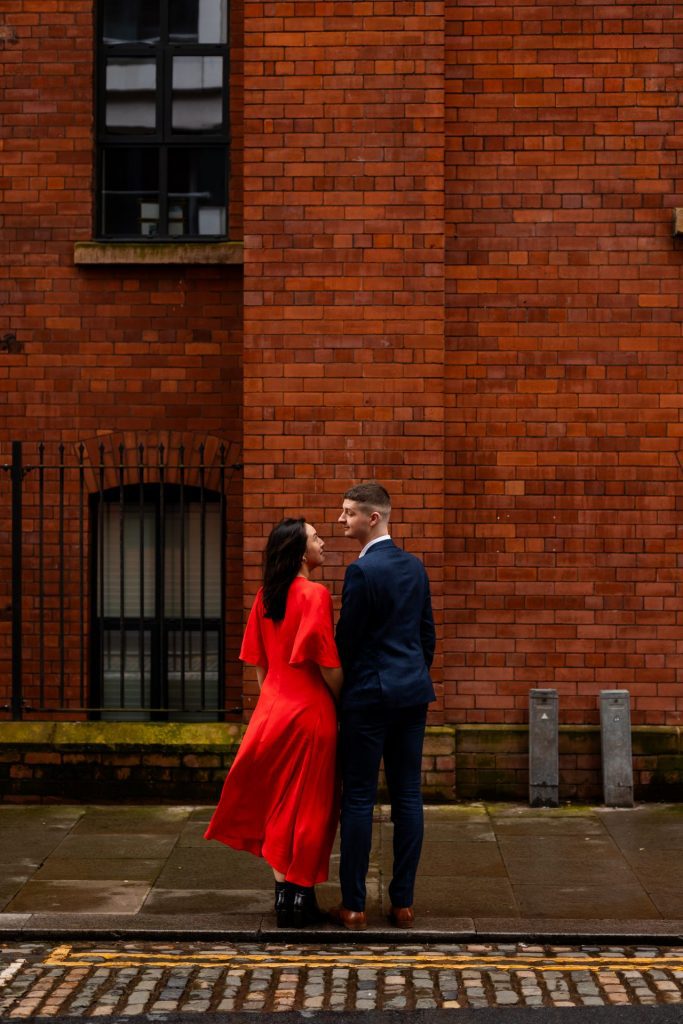 Newly engaged couple walking together in Ancoats, Manchester - The bride to-be is wearing a vivid red dress & the groom to-be is wearing a blue suit - Engagement Shoot - Isabelle B Photography