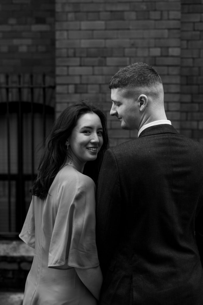 Newly engaged couple walking together in Ancoats, Manchester - The bride to-be is wearing a vivid red dress & the groom to-be is wearing a blue suit - Engagement Shoot - Black & White - Isabelle B Photography