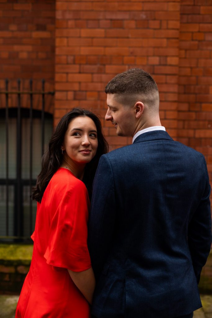 Newly engaged couple walking together in Ancoats, Manchester - The bride to-be is wearing a vivid red dress & the groom to-be is wearing a blue suit - Engagement Shoot - Isabelle B Photography