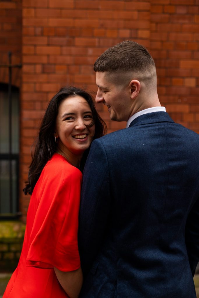 Newly engaged couple walking together in Ancoats, Manchester - The bride to-be is wearing a vivid red dress & the groom to-be is wearing a blue suit - Engagement Shoot - Isabelle B Photography