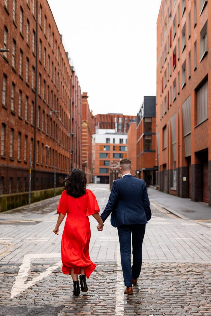 Newly engaged couple walking together in Ancoats, Manchester - The bride to-be is wearing a vivid red dress & the groom to-be is wearing a blue suit - Engagement Shoot - Isabelle B Photography