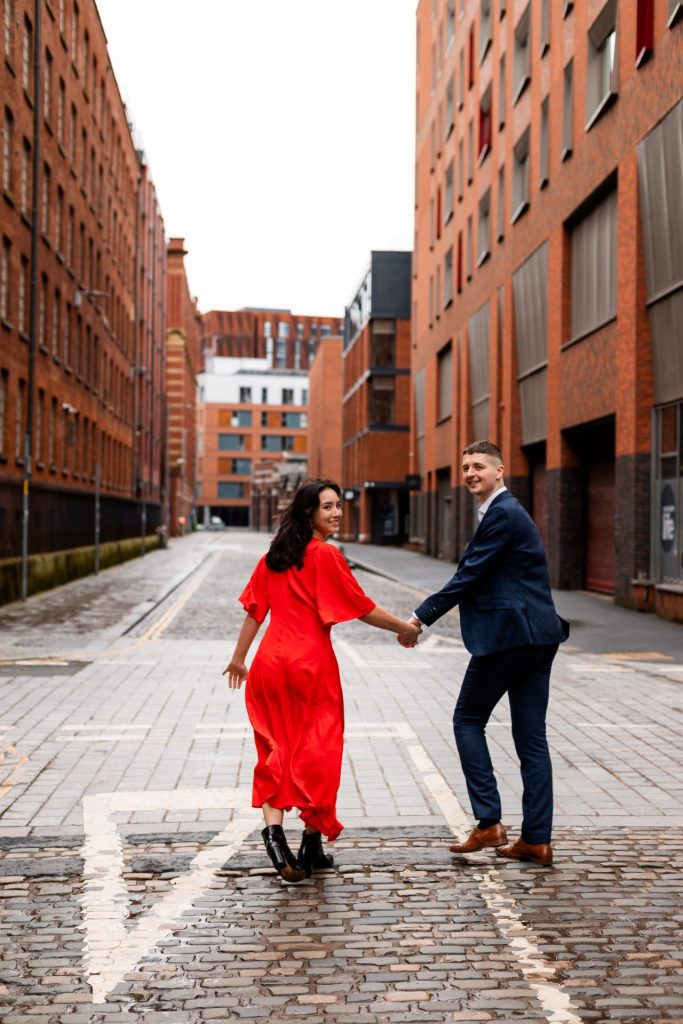 Newly engaged couple walking together in Ancoats, Manchester - The bride to-be is wearing a vivid red dress & the groom to-be is wearing a blue suit - Engagement Shoot - Isabelle B Photography