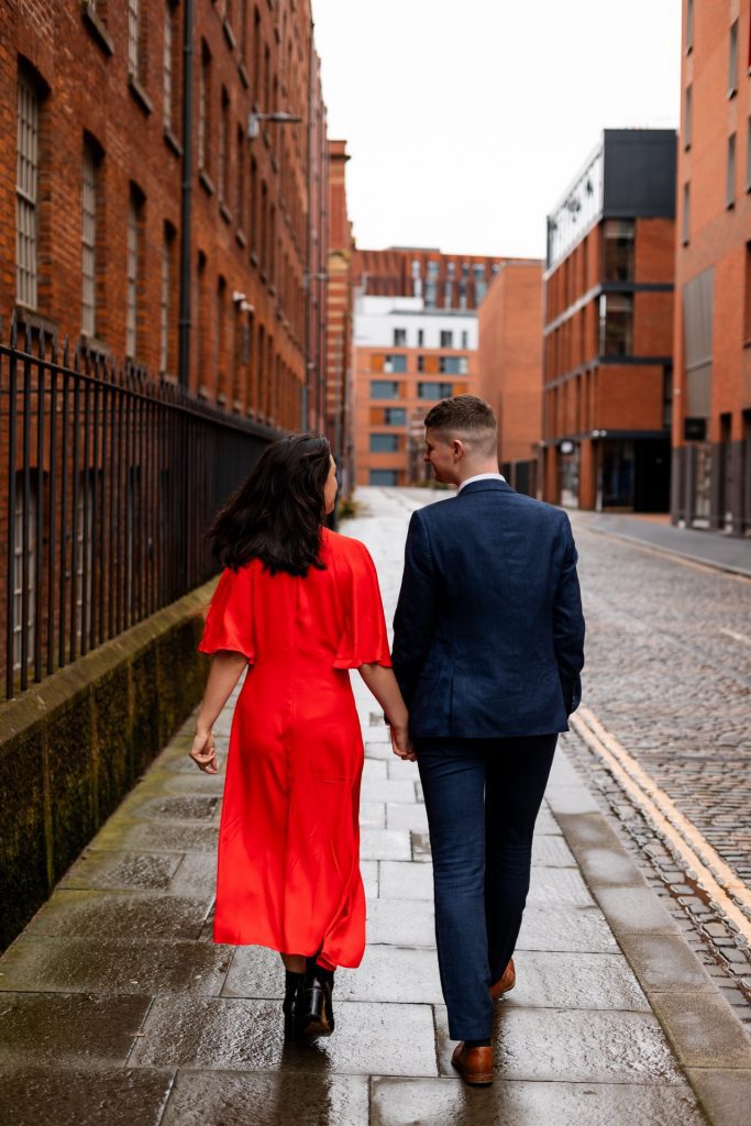 Newly engaged couple walking together in Ancoats, Manchester - The bride to-be is wearing a vivid red dress & the groom to-be is wearing a blue suit - Engagement Shoot - Isabelle B Photography