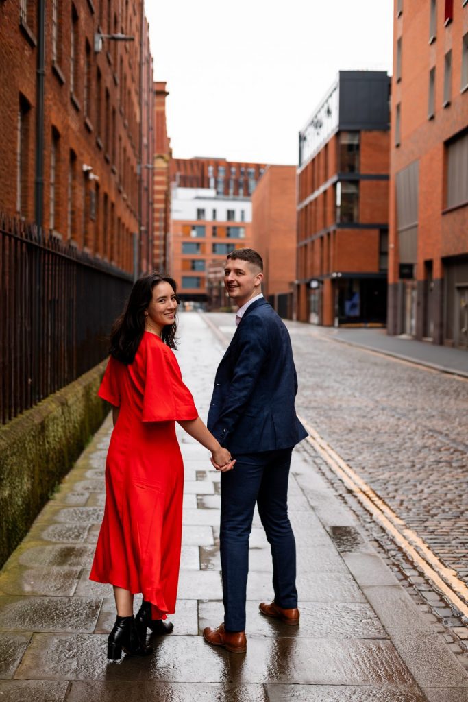 Newly engaged couple walking together in Ancoats, Manchester - The bride to-be is wearing a vivid red dress & the groom to-be is wearing a blue suit - Engagement Shoot - Isabelle B Photography
