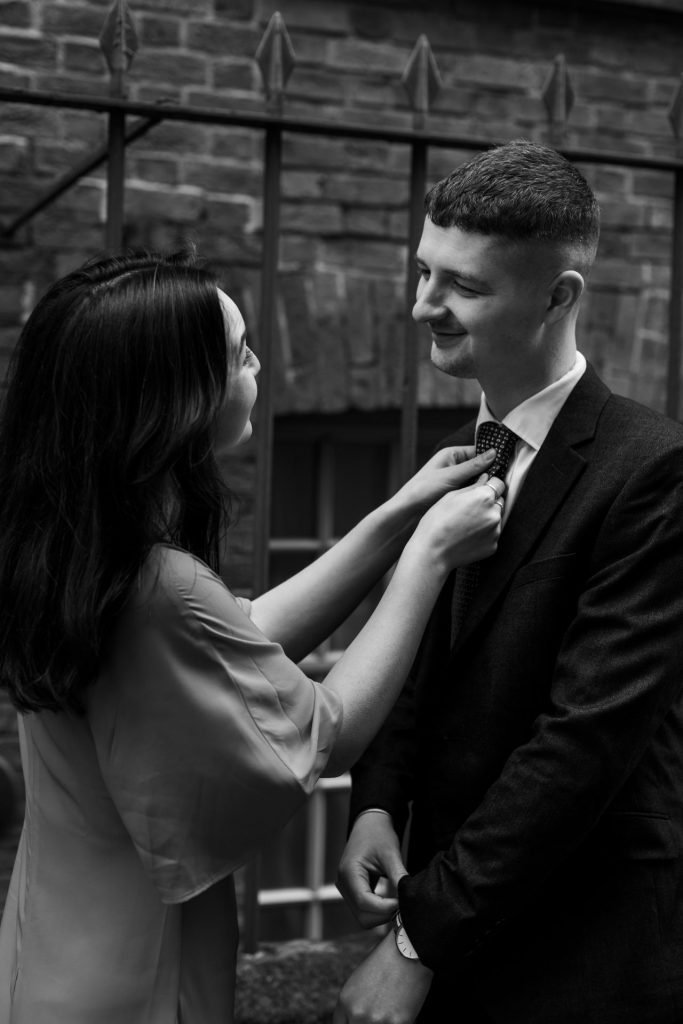 Newly engaged couple walking together in Ancoats, Manchester - The bride to-be is wearing a vivid red dress & the groom to-be is wearing a blue suit - The bride is adjusting your fiancés tie - Engagement Shoot - Black & White - Isabelle B Photography