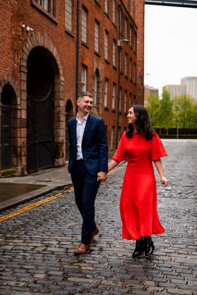 Newly engaged couple walking together in Ancoats, Manchester - The bride to-be is wearing a vivid red dress & the groom to-be is wearing a blue suit - Engagement Shoot - Isabelle B Photography