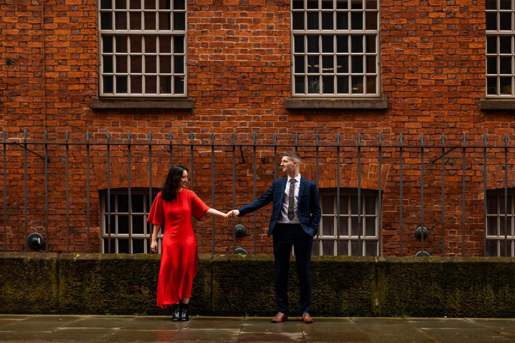 Newly engaged couple walking together in Ancoats, Manchester - The bride to-be is wearing a vivid red dress & the groom to-be is wearing a blue suit - Engagement Shoot - Isabelle B Photography