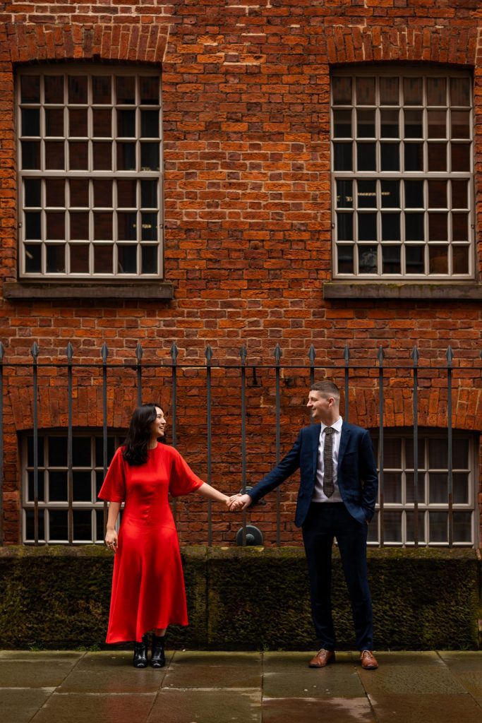Newly engaged couple walking together in Ancoats, Manchester - The bride to-be is wearing a vivid red dress & the groom to-be is wearing a blue suit - Engagement Shoot - Isabelle B Photography
