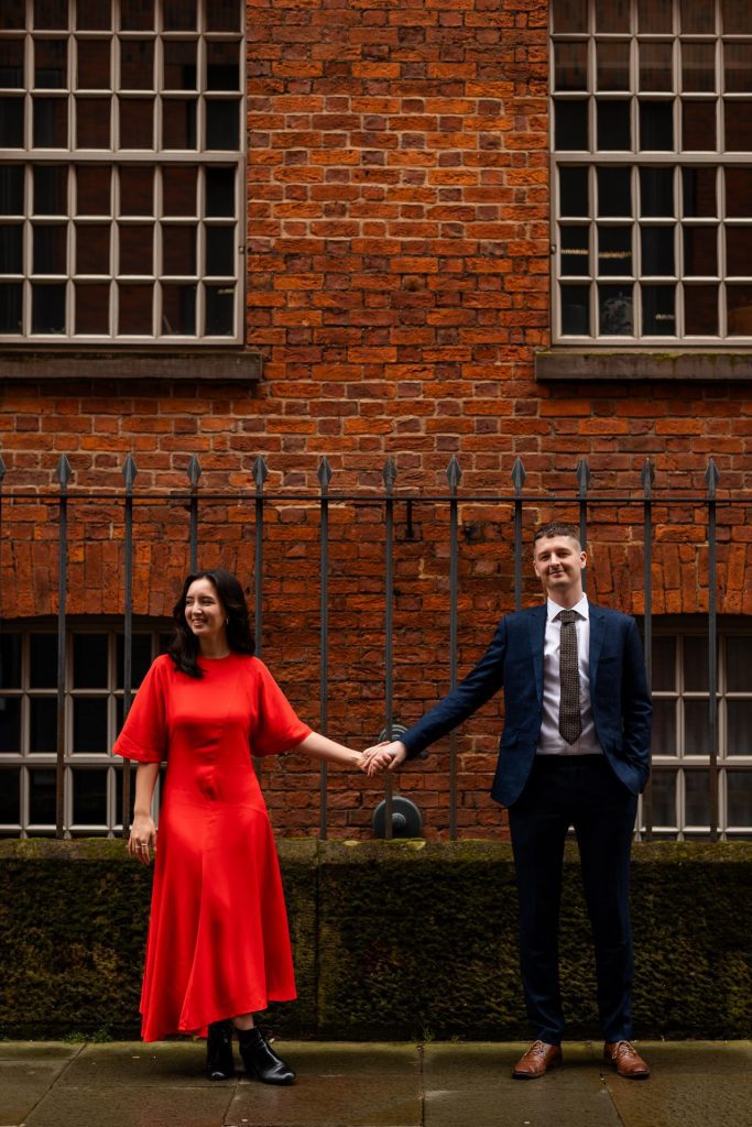 Newly engaged couple walking together in Ancoats, Manchester - The bride to-be is wearing a vivid red dress & the groom to-be is wearing a blue suit - Engagement Shoot - Isabelle B Photography