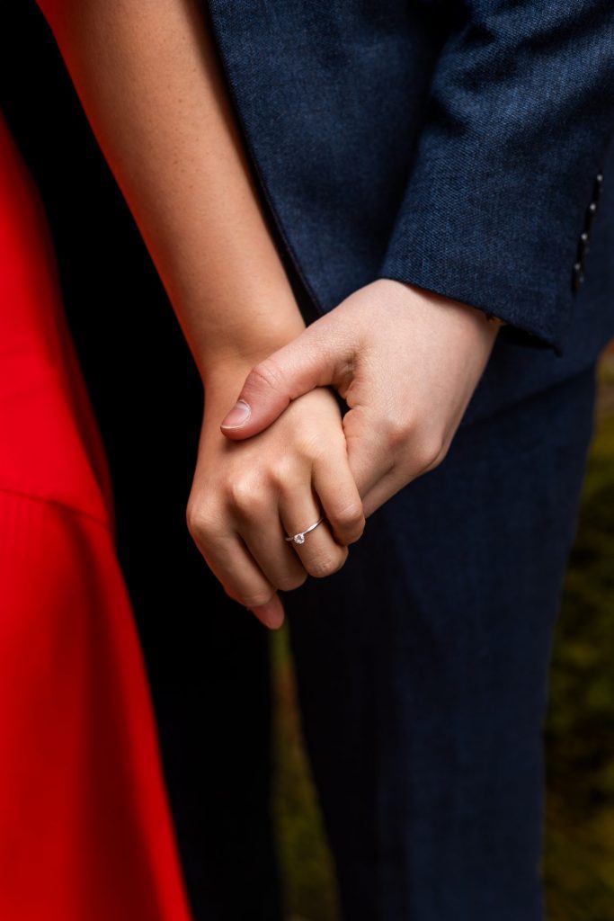 Close up of engagement ring on the bride to-be's hand again a red rick wall in Ancoats, Manchester - The bride to-be is wearing a vivid red dress & the groom to-be is wearing a blue suit - Engagement Shoot - Isabelle B Photography