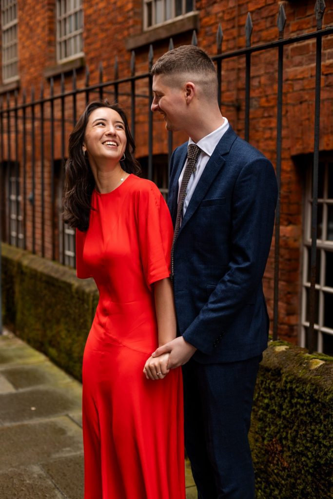 Newly engaged couple walking together in Ancoats, Manchester - The bride to-be is wearing a vivid red dress & the groom to-be is wearing a blue suit - Engagement Shoot - Isabelle B Photography