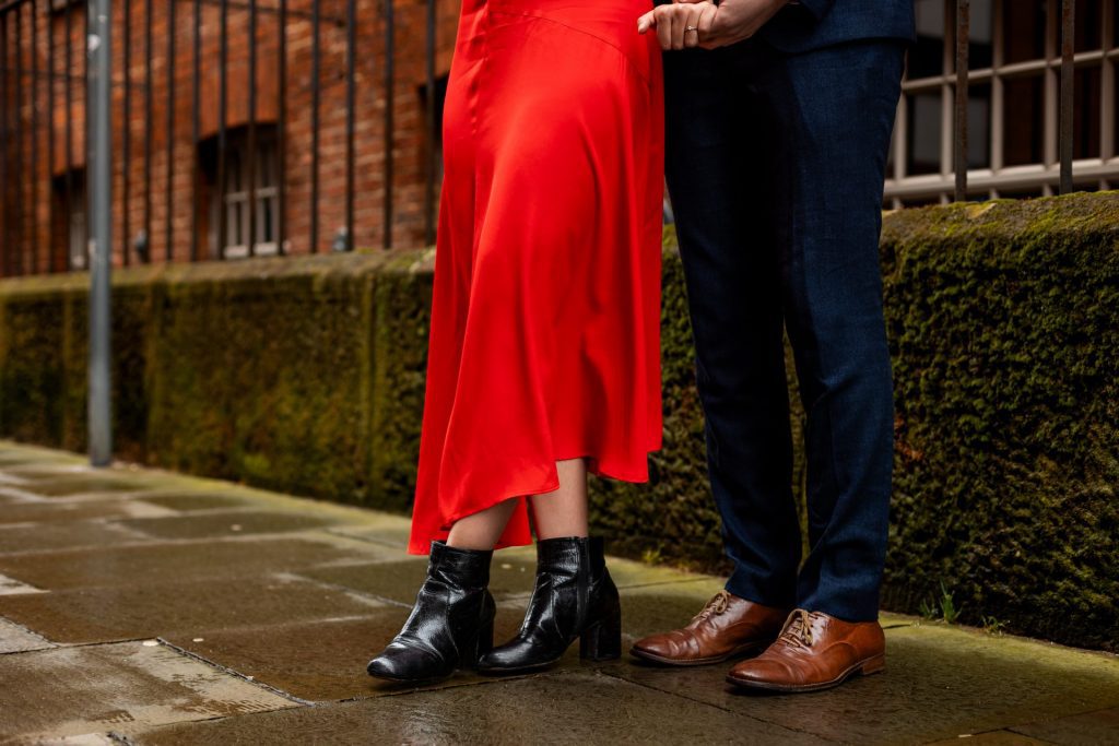 Close up of the couples shoes against a red rick wall in Ancoats, Manchester - The bride to-be is wearing a vivid red dress & the groom to-be is wearing a blue suit - Engagement Shoot - Isabelle B Photography