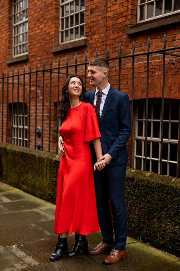 Newly engaged couple walking together in Ancoats, Manchester - The bride to-be is wearing a vivid red dress & the groom to-be is wearing a blue suit - Engagement Shoot - Isabelle B Photography