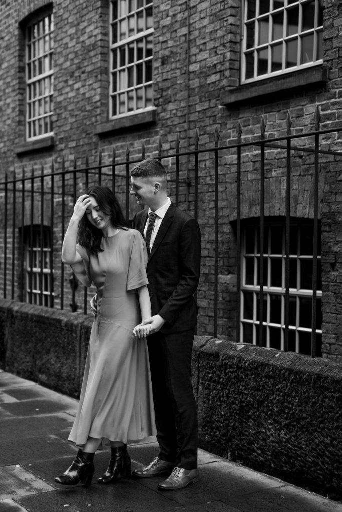 Newly engaged couple walking together in Ancoats, Manchester - The bride to-be is wearing a vivid red dress & the groom to-be is wearing a blue suit - Engagement Shoot - Black & White - Isabelle B Photography