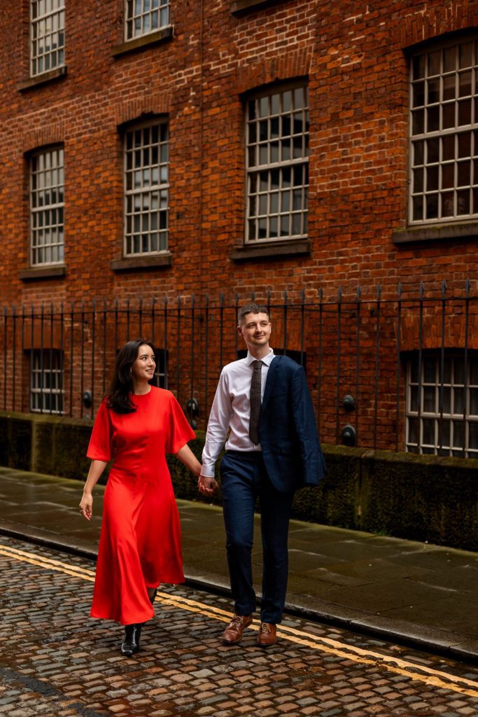 Newly engaged couple walking together in Ancoats, Manchester - The bride to-be is wearing a vivid red dress & the groom to-be is wearing a blue suit - Engagement Shoot - Isabelle B Photography