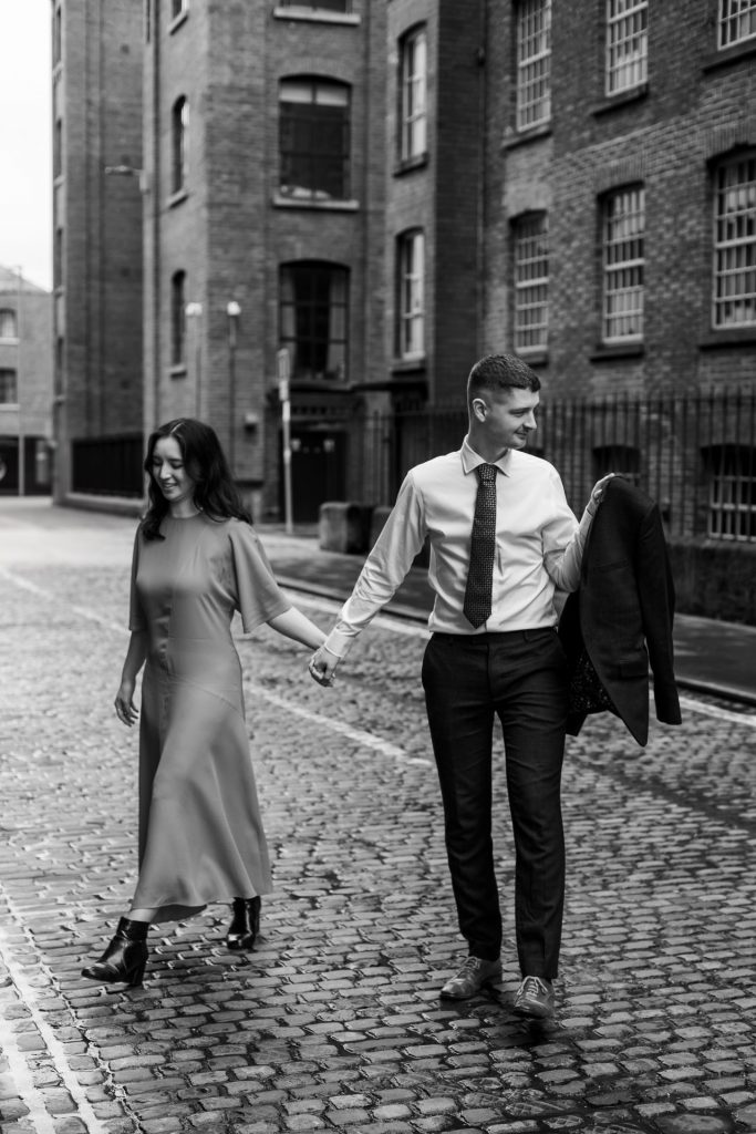 Newly engaged couple walking together in Ancoats, Manchester - The bride to-be is wearing a vivid red dress & the groom to-be is wearing a blue suit - Engagement Shoot - Black & White - Isabelle B Photography
