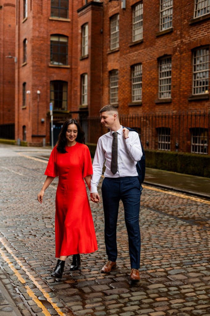 Newly engaged couple walking together in Ancoats, Manchester - The bride to-be is wearing a vivid red dress & the groom to-be is wearing a blue suit - Engagement Shoot - Isabelle B Photography