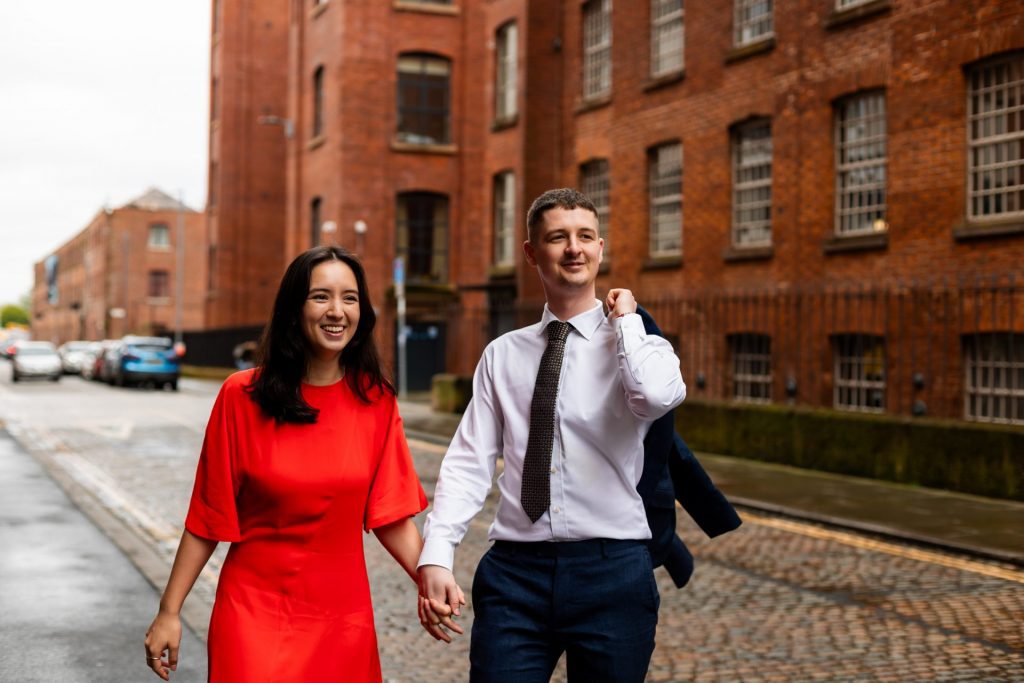 Newly engaged couple walking together in Ancoats, Manchester - The bride to-be is wearing a vivid red dress & the groom to-be is wearing a blue suit - Engagement Shoot - Isabelle B Photography
