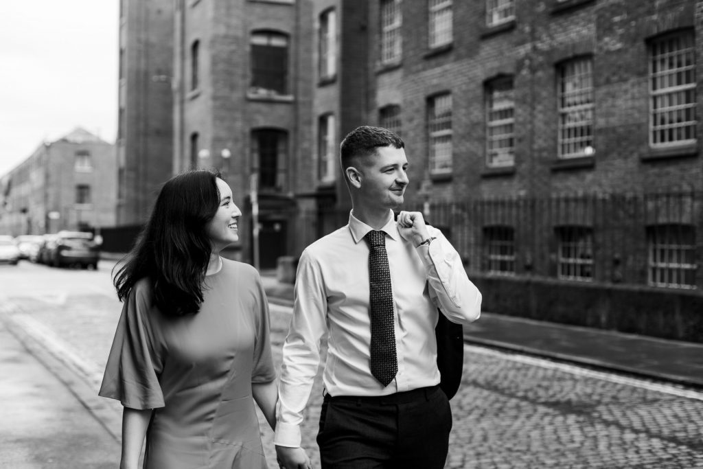 Newly engaged couple walking together in Ancoats, Manchester - The bride to-be is wearing a vivid red dress & the groom to-be is wearing a blue suit - Engagement Shoot - Black & White - Isabelle B Photography