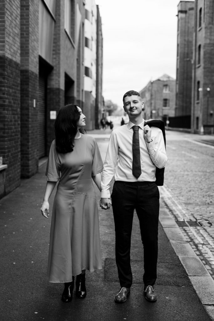 Newly engaged couple walking together in Ancoats, Manchester - The bride to-be is wearing a vivid red dress & the groom to-be is wearing a blue suit - Engagement Shoot - Black & White - Isabelle B Photography