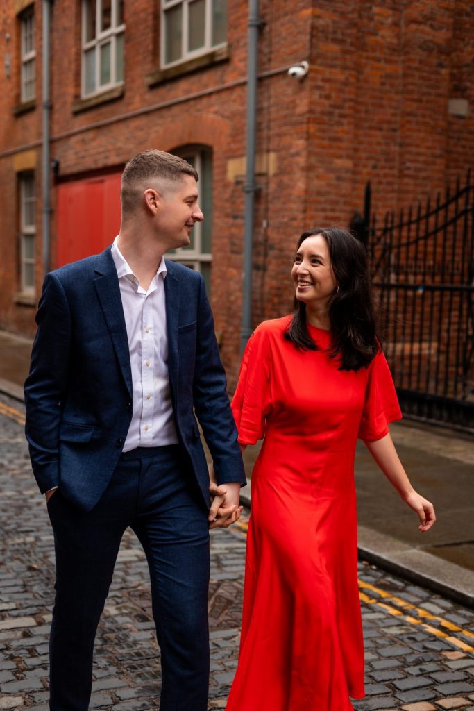 Newly engaged couple walking together in Ancoats, Manchester - The bride to-be is wearing a vivid red dress & the groom to-be is wearing a blue suit - Engagement Shoot - Isabelle B Photography
