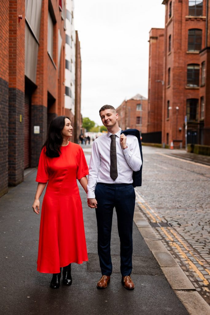 Newly engaged couple walking together in Ancoats, Manchester - The bride to-be is wearing a vivid red dress & the groom to-be is wearing a blue suit - Engagement Shoot - Isabelle B Photography