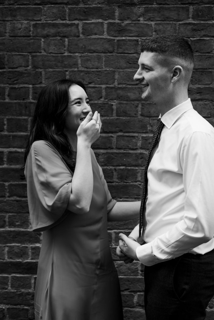 Newly engaged couple walking together in Ancoats, Manchester - The bride to-be is wearing a vivid red dress & the groom to-be is wearing a blue suit - Engagement Shoot - Black & White - Isabelle B Photography