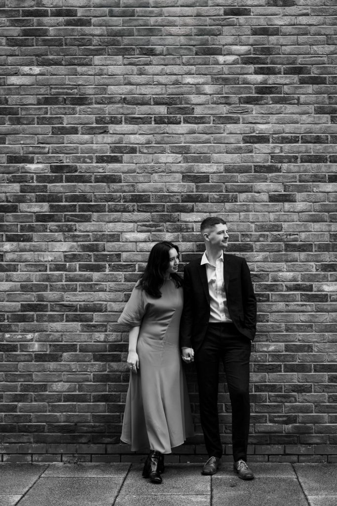 Newly engaged couple walking together in Ancoats, Manchester - The bride to-be is wearing a vivid red dress & the groom to-be is wearing a blue suit - Engagement Shoot - Black & White Isabelle B Photography